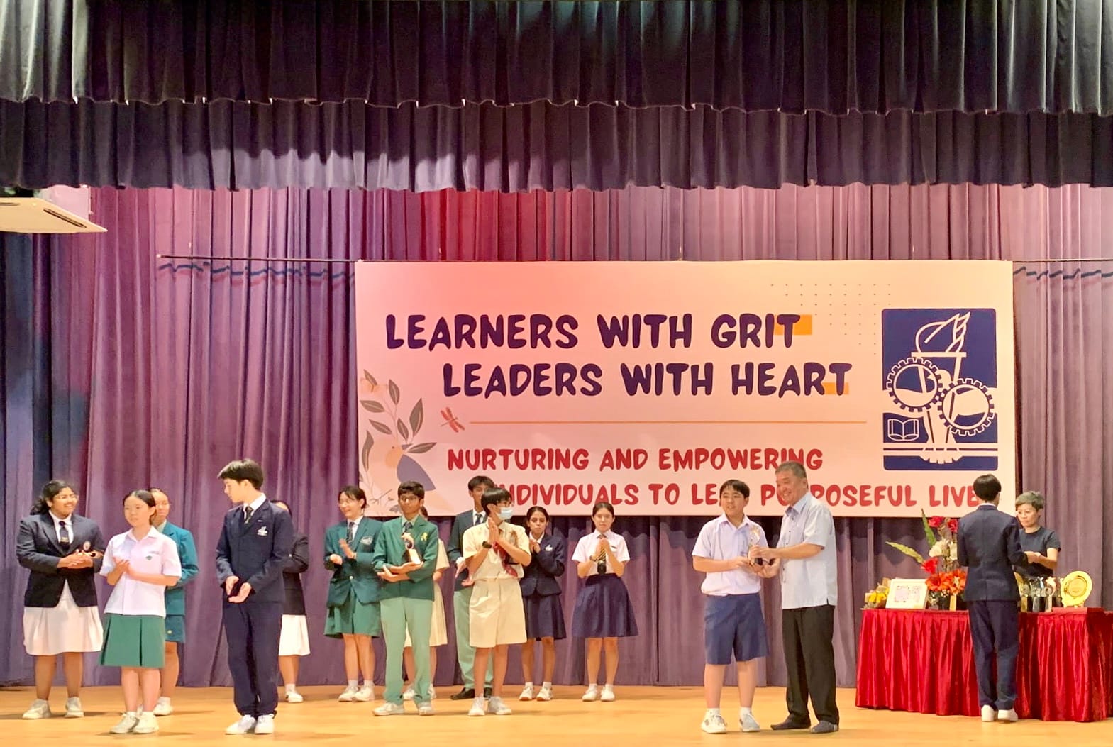 Students in uniforms stand on a stage receiving awards in front of a banner reading Learners With Grit Leaders With Heart.